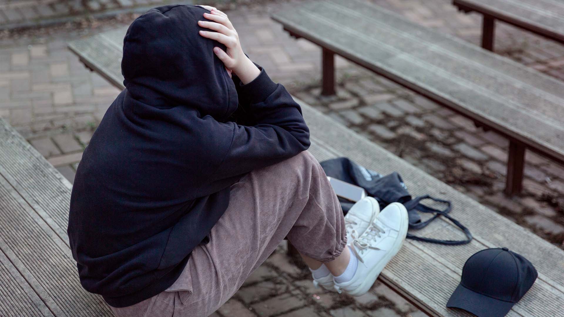 Teenager sidder på bænk med ryggen til kameraet og ansigtet skjult i sine hænder. FOTO: AdobeStock