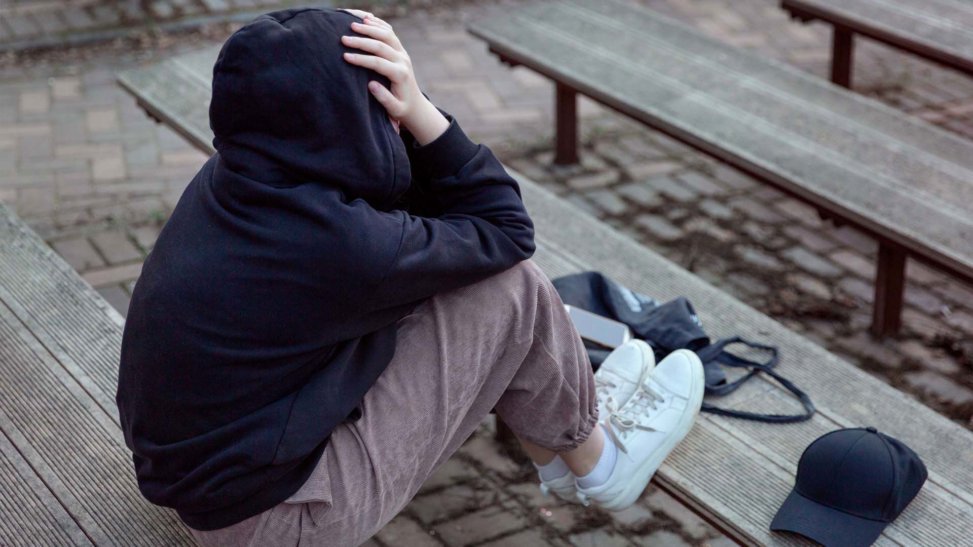 Teenager sidder på bænk med ryggen til kameraet og ansigtet skjult i sine hænder. FOTO: AdobeStock